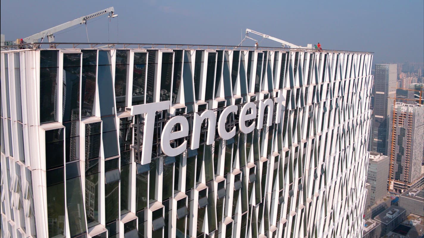 Tencent logo in large white letters on a glass office tower’s facade, with roof cranes and a city skyline behind