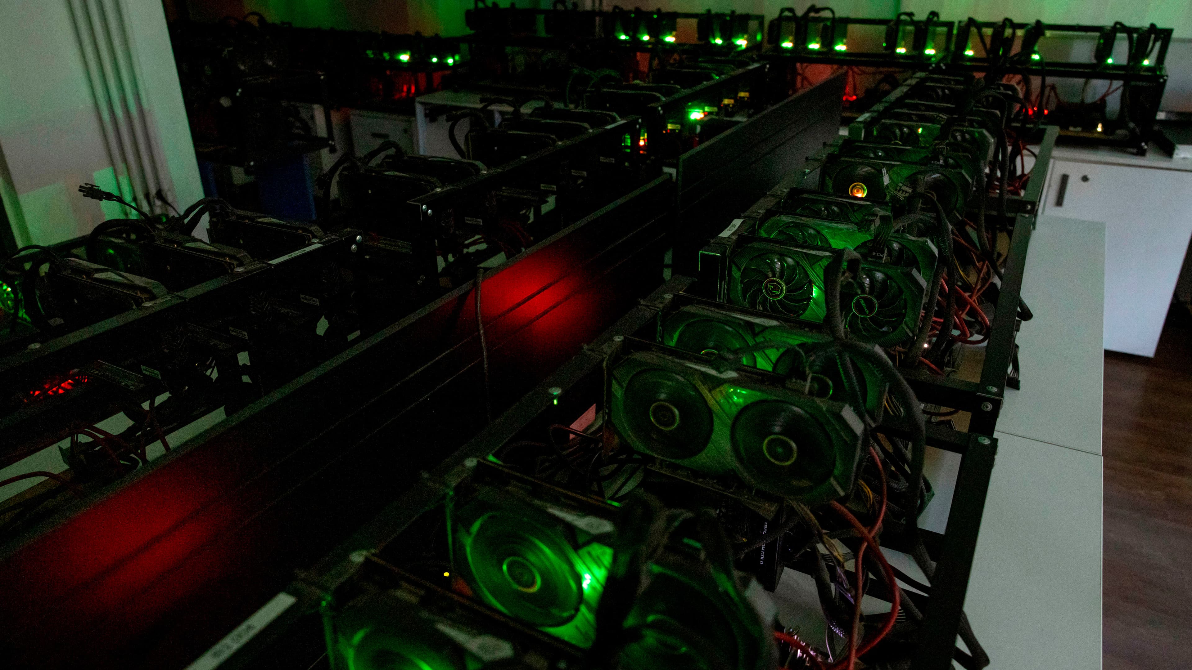 Rows of open-frame cryptocurrency mining rigs packed with graphics cards, their fans glowing green, in a dimly lit room