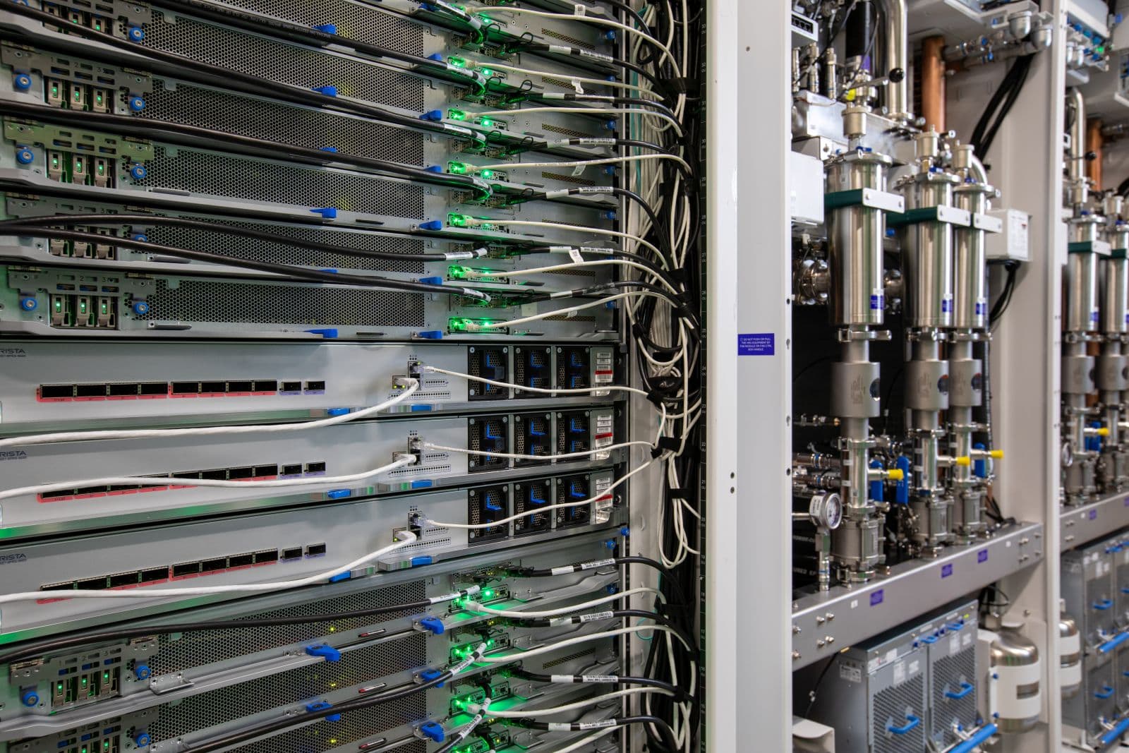 Close-up of server racks with connected cables and green indicator lights beside metal piping and cooling equipment.
