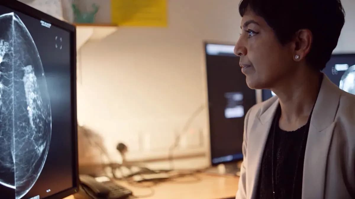 A healthcare professional with short dark hair and a light jacket looking at a mammogram scan displayed on a computer screen.